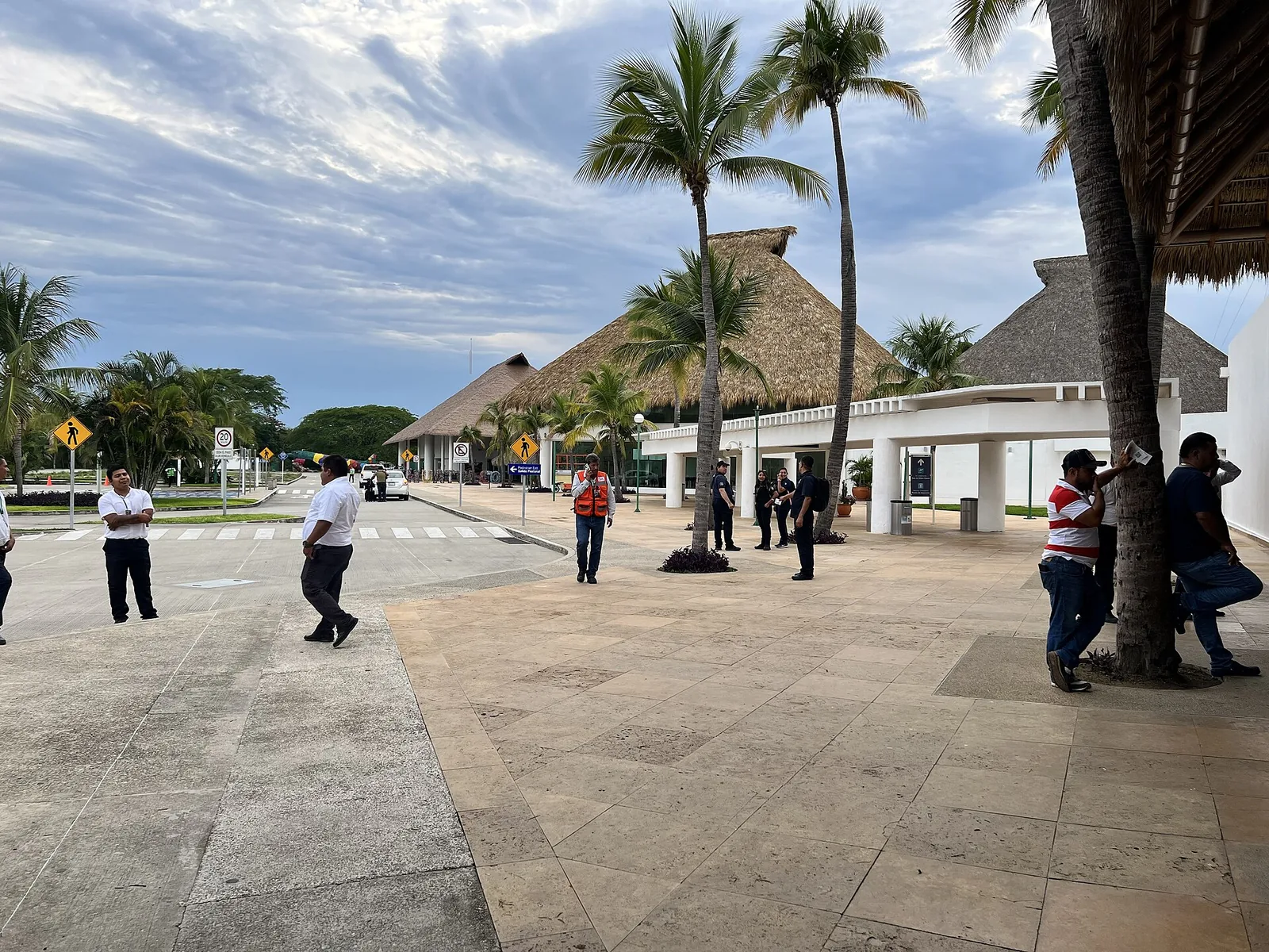Acceso del aeropuerto de Huatulco, con techos de palapa sobre la terminal, palmeras altas y personal esperando en la acera.