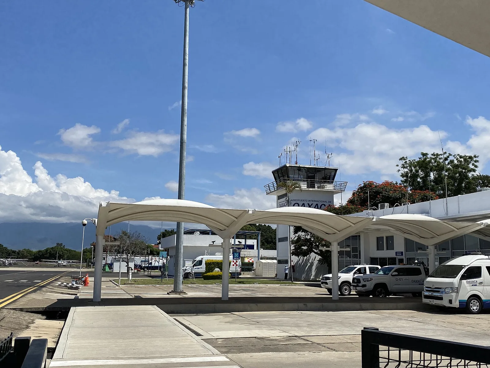Zona de recogida del aeropuerto de Oaxaca, con marquesinas blancas, camionetas de transporte esperando y la torre de control al fondo.