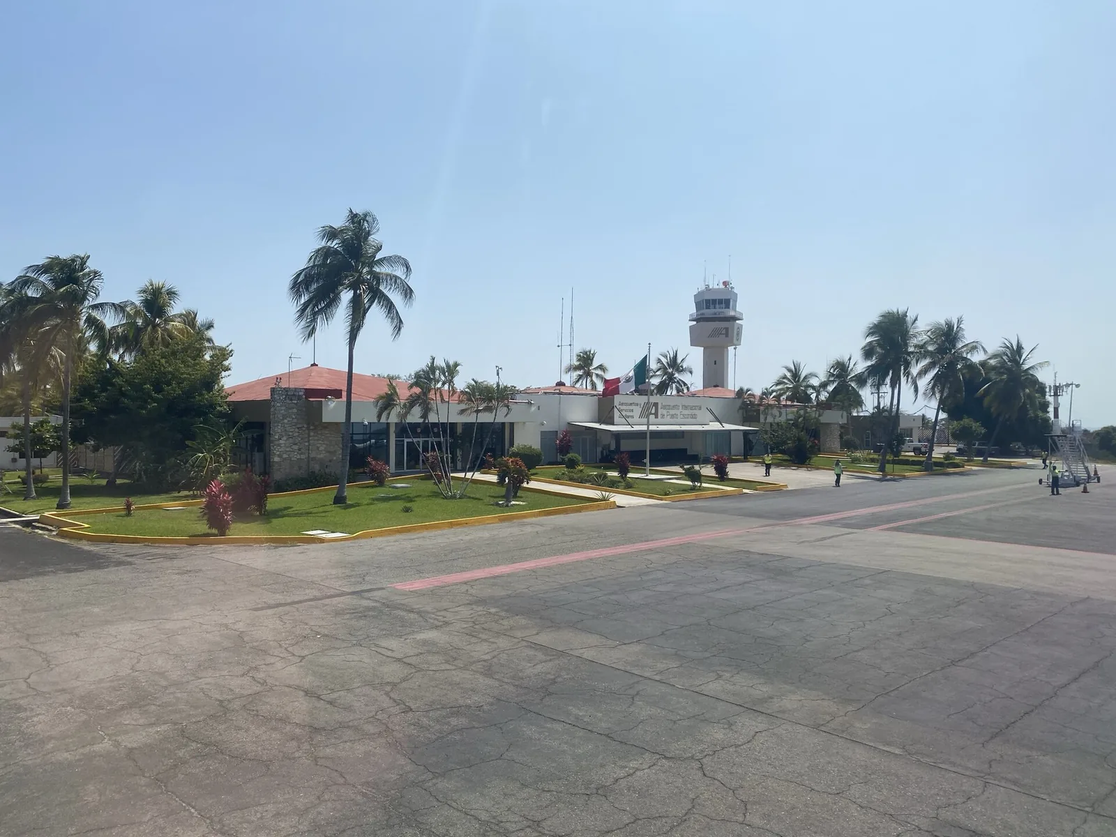 Exterior del aeropuerto de Puerto Escondido: la torre de control entre palmeras y el edificio de la terminal al fondo del acceso.