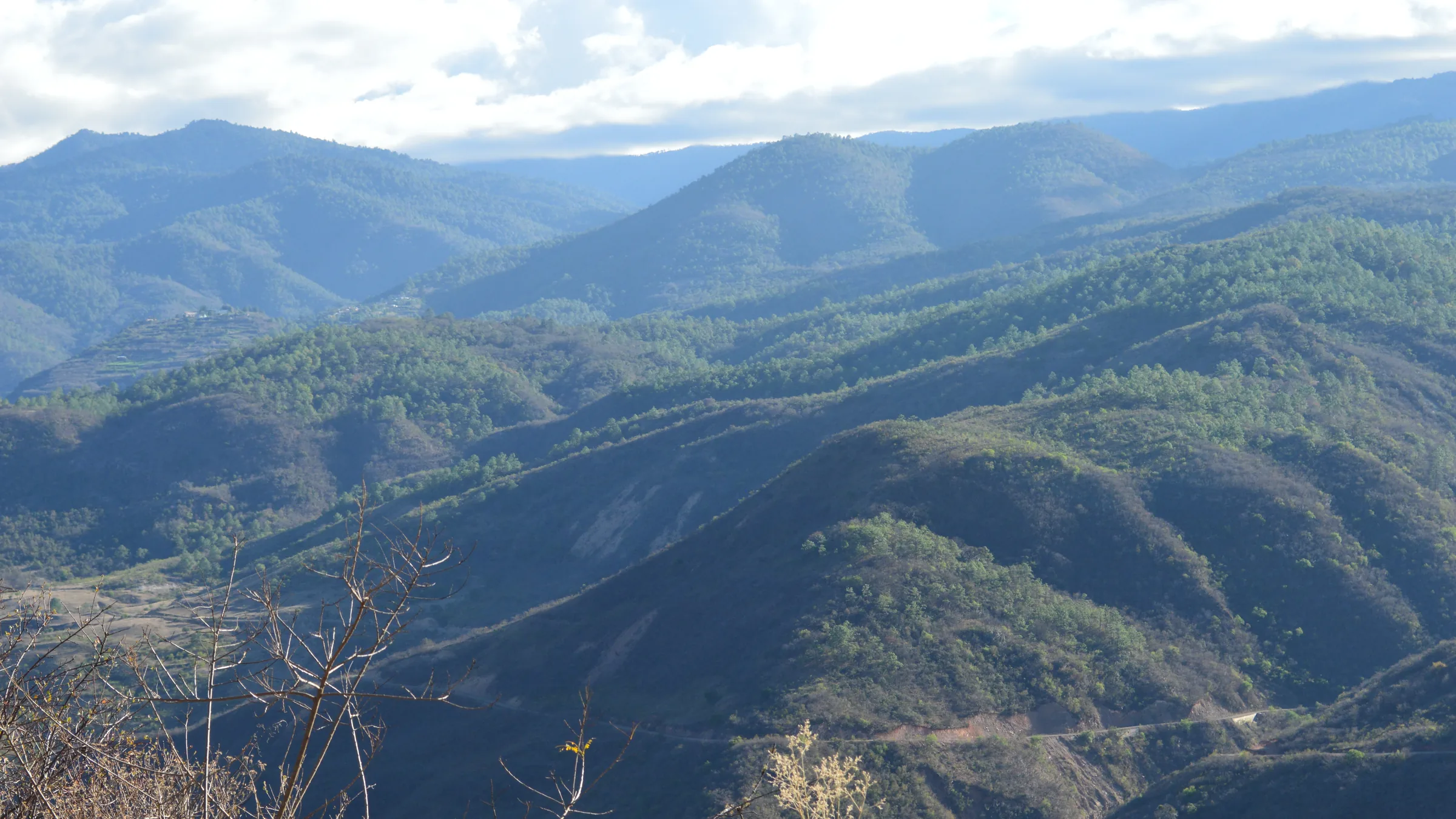 Cerros boscosos de la sierra de Oaxaca uno detrás de otro bajo un cielo nublado, con la cinta de la carretera federal 175 recorriendo la ladera del cerro más cercano.