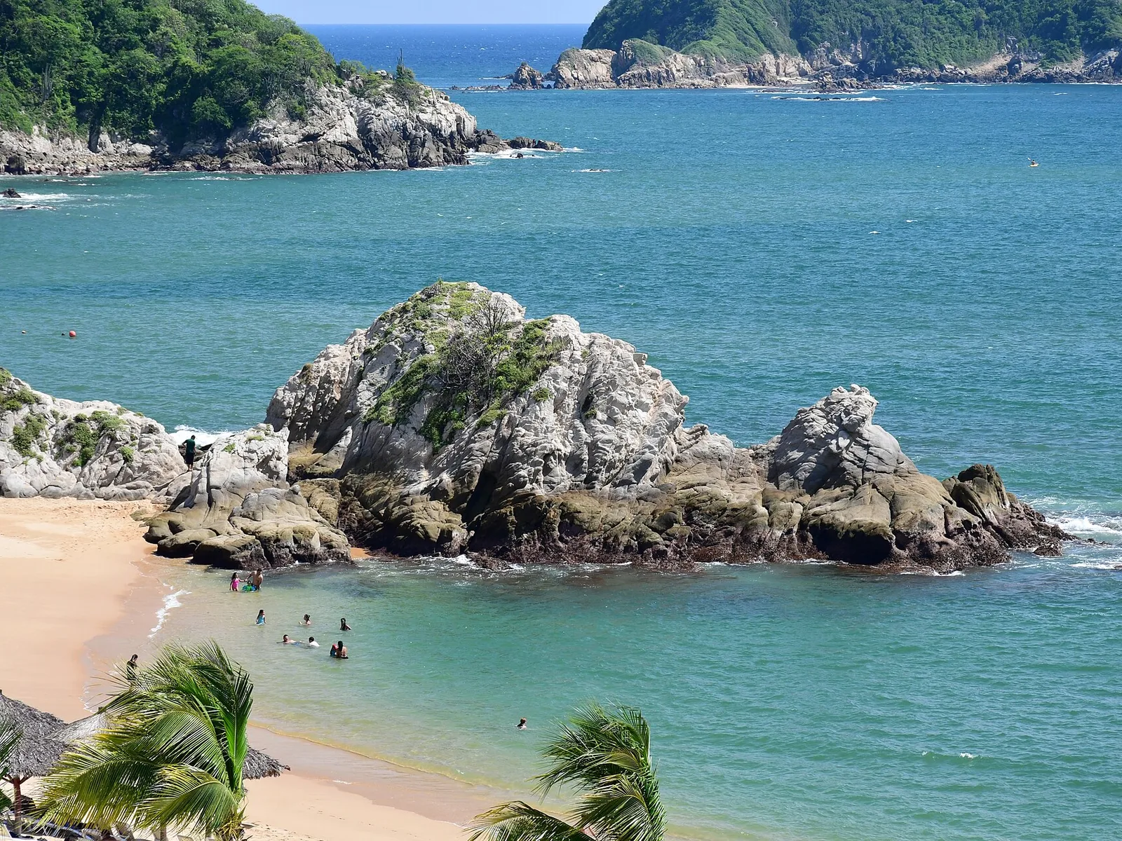 Bahía de Huatulco con formaciones rocosas dentro del agua, arena clara y vegetación en el acantilado.