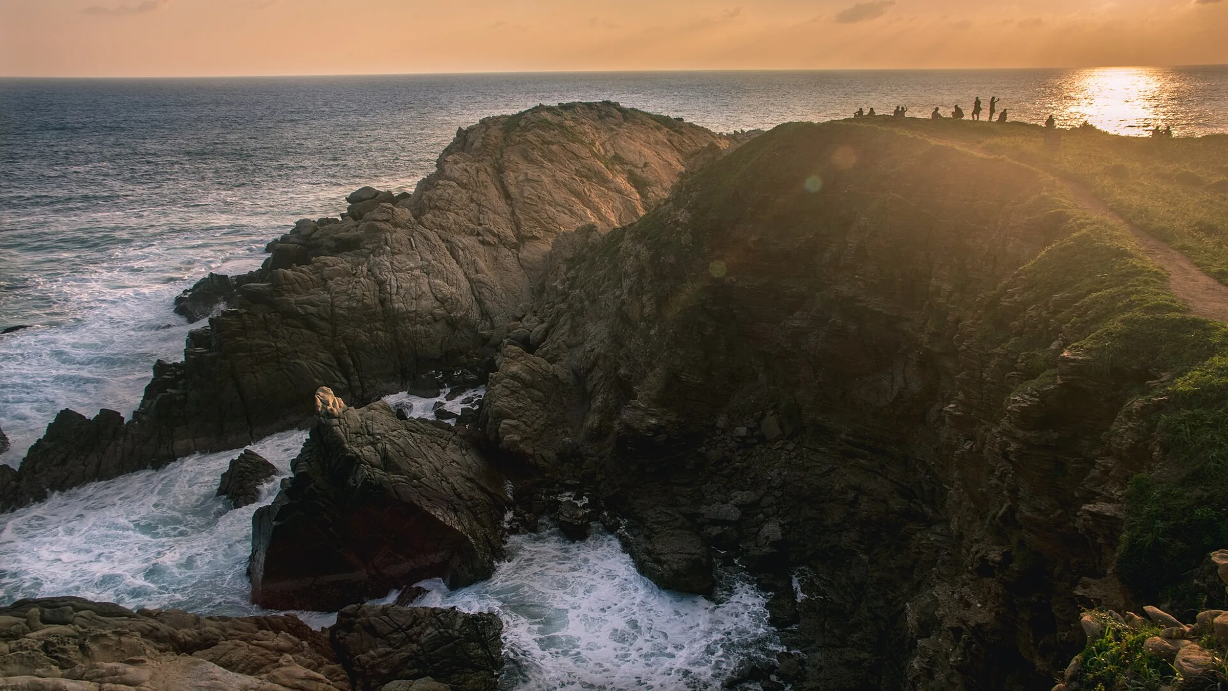 Punta Cometa al atardecer: el promontorio rocoso de Mazunte metiéndose en el mar, las olas rompiendo contra la roca y unas pocas siluetas de personas en lo alto del cerro recortadas contra el sol bajo.