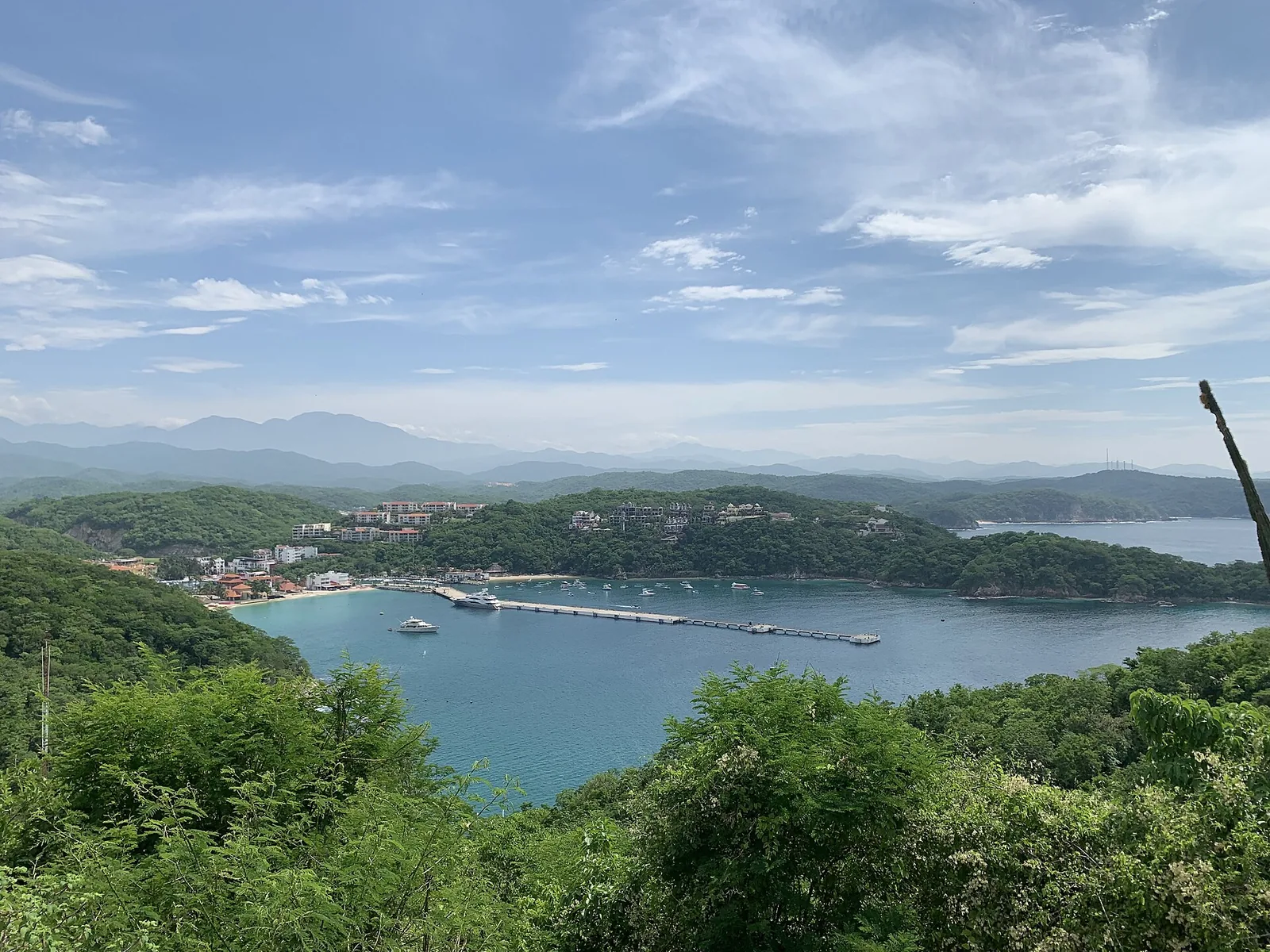 Vista de la bahía de Santa Cruz en Huatulco desde el mirador: el muelle de cruceros adentrándose en el agua turquesa, yates fondeados, el pueblo con sus tejados rojos en la orilla y los cerros verdes cerrando la bahía.