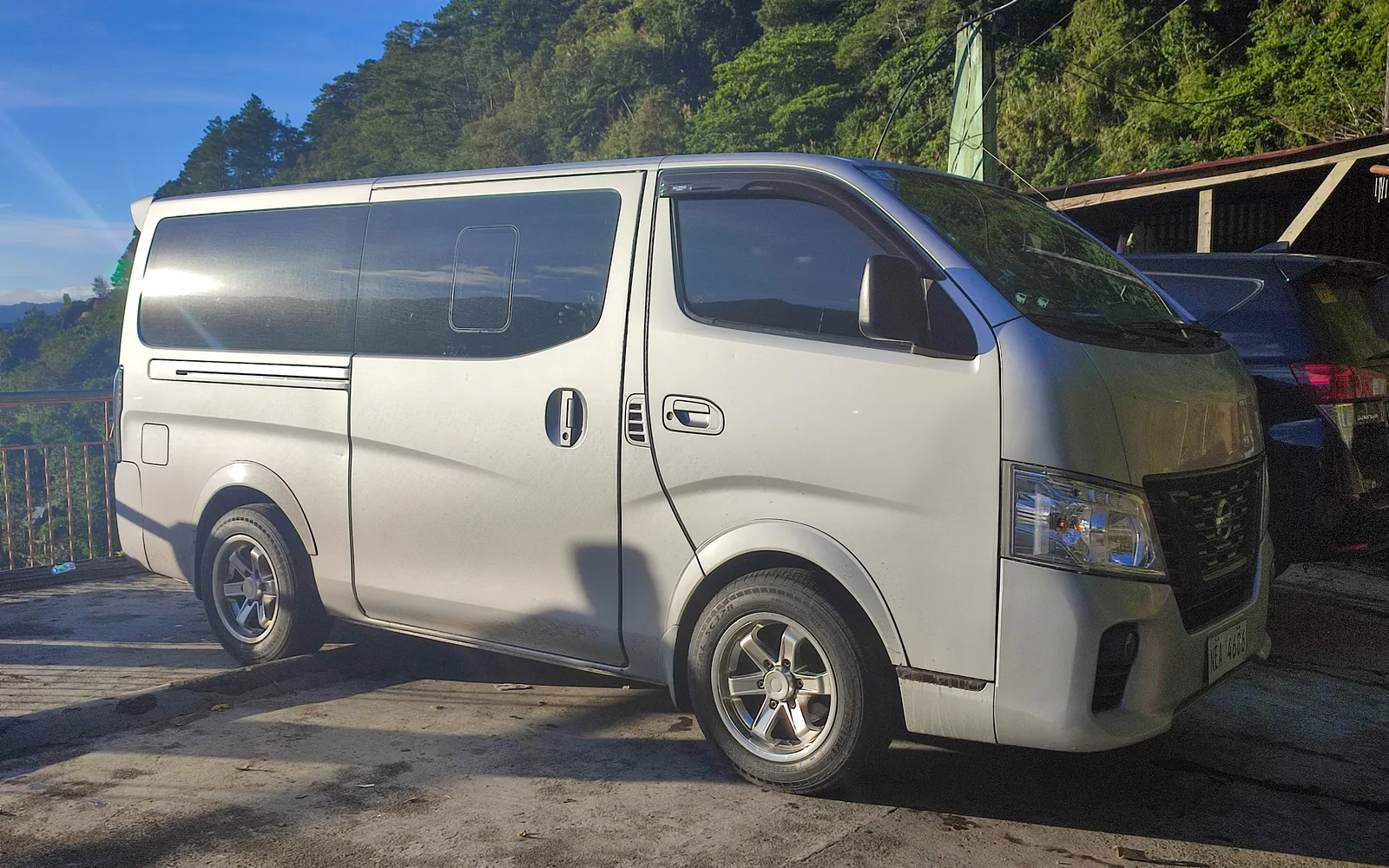 Una van Nissan NV350 Urvan plateada aparcada en una carretera de montaña, vista de lado y tres cuartos delantera, con cielo azul y una ladera boscosa detrás.