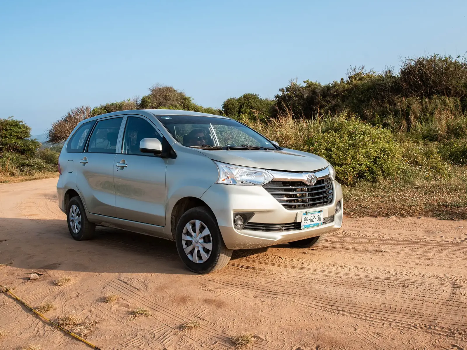 Un Toyota Avanza plateado de Eco Adventures sobre un camino de arena junto a la costa, visto de frente y tres cuartos, con matorral bajo detrás.