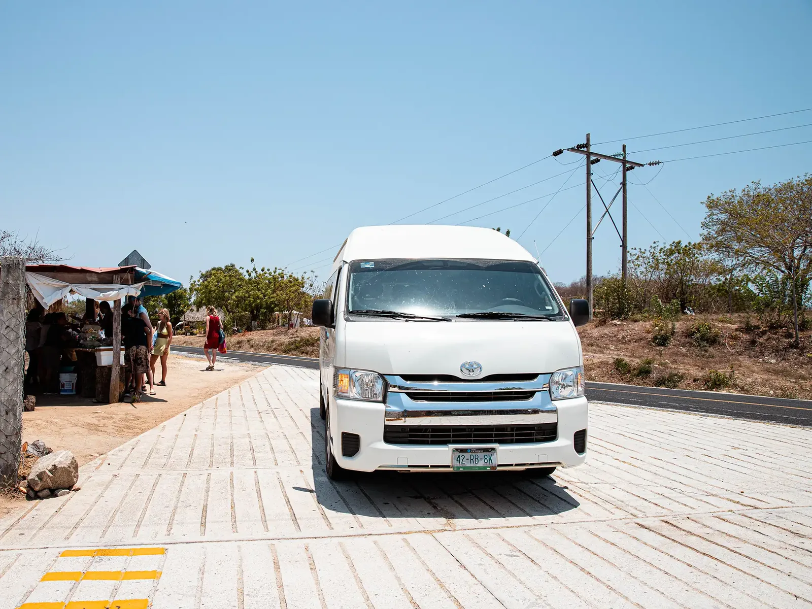 Una van Toyota Hiace blanca de Eco Adventures parada en la carretera de la costa, vista de frente y tres cuartos, con el mar y la vegetación seca al fondo y un puesto de comida a un lado del camino.