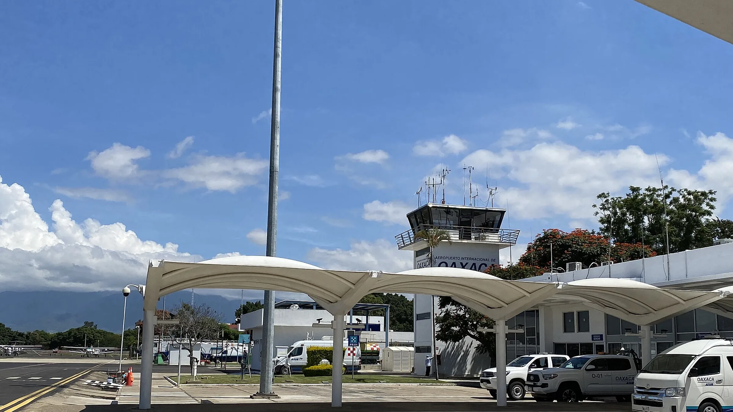 Salida de llegadas del aeropuerto de Oaxaca: marquesinas blancas sobre la acera, camionetas y taxis oficiales del módulo de transporte esperando, y la torre de control con el letrero del aeropuerto al fondo.