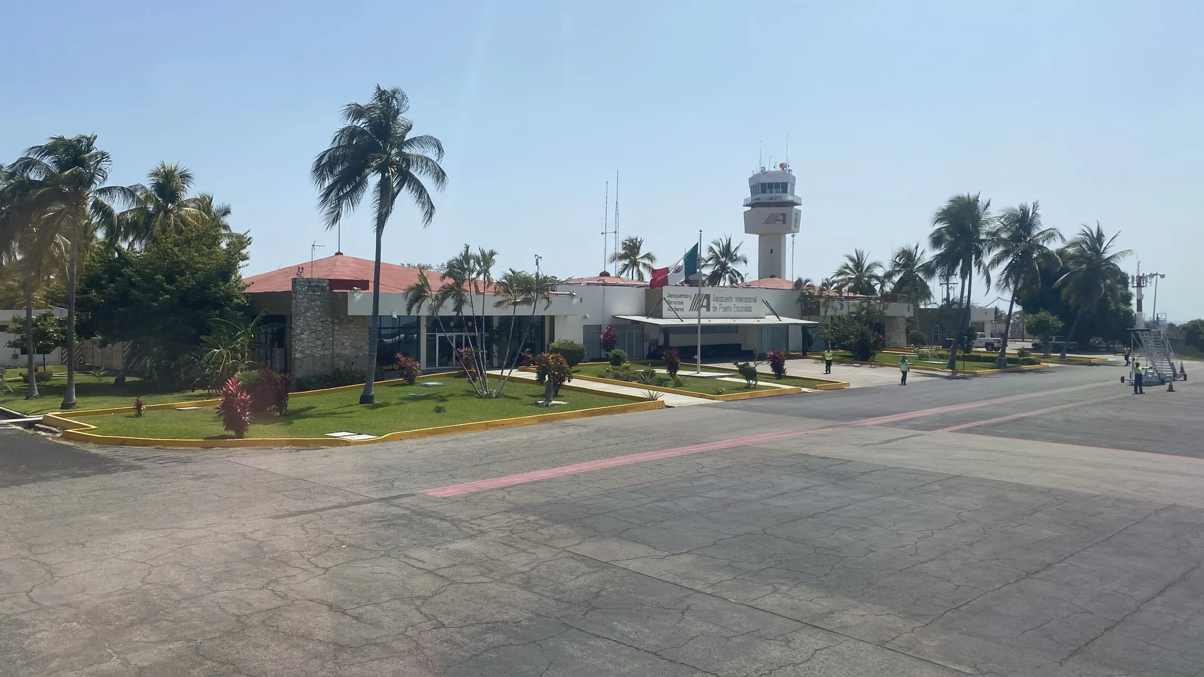 Exterior del aeropuerto de Puerto Escondido: la torre de control asomando entre palmeras, el edificio bajo de la terminal con su tejado rojo y la plataforma de asfalto en primer plano.