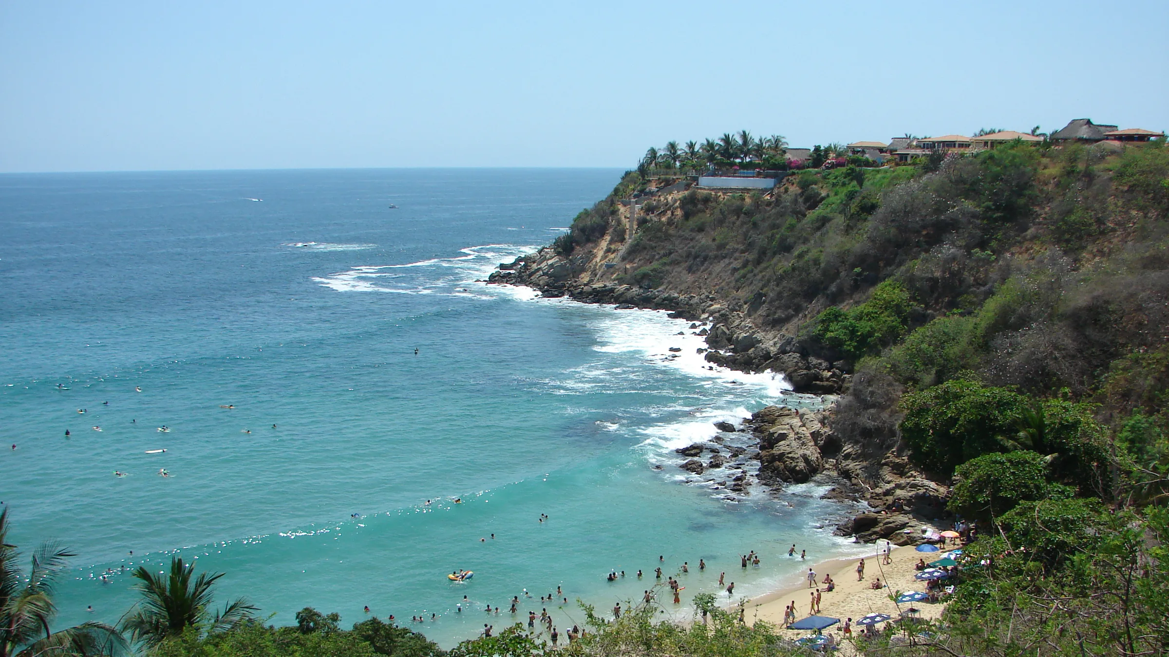 La cala de Carrizalillo vista desde el acantilado: el agua turquesa y en calma dentro de la herradura, bañistas y tablas de surf cerca de la orilla, la franja de arena con sombrillas al fondo y, arriba del acantilado, las casas y palmeras del bulevar de Rinconada.