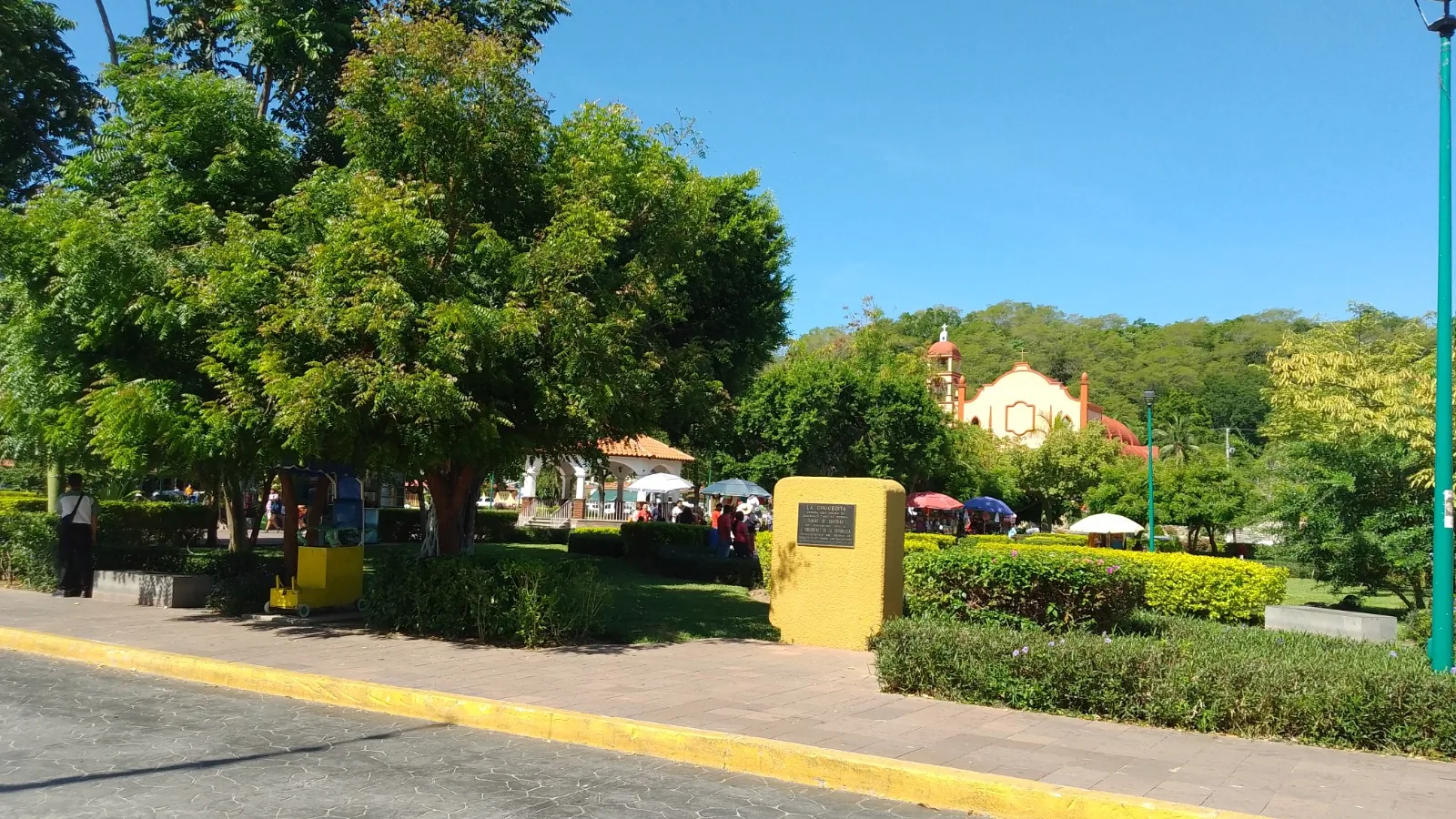 La plaza de La Crucecita, en Huatulco: árboles grandes dando sombra sobre el césped, puestos con sombrillas de colores al fondo y la iglesia de fachada rosada con su campanario recortada contra el cerro.