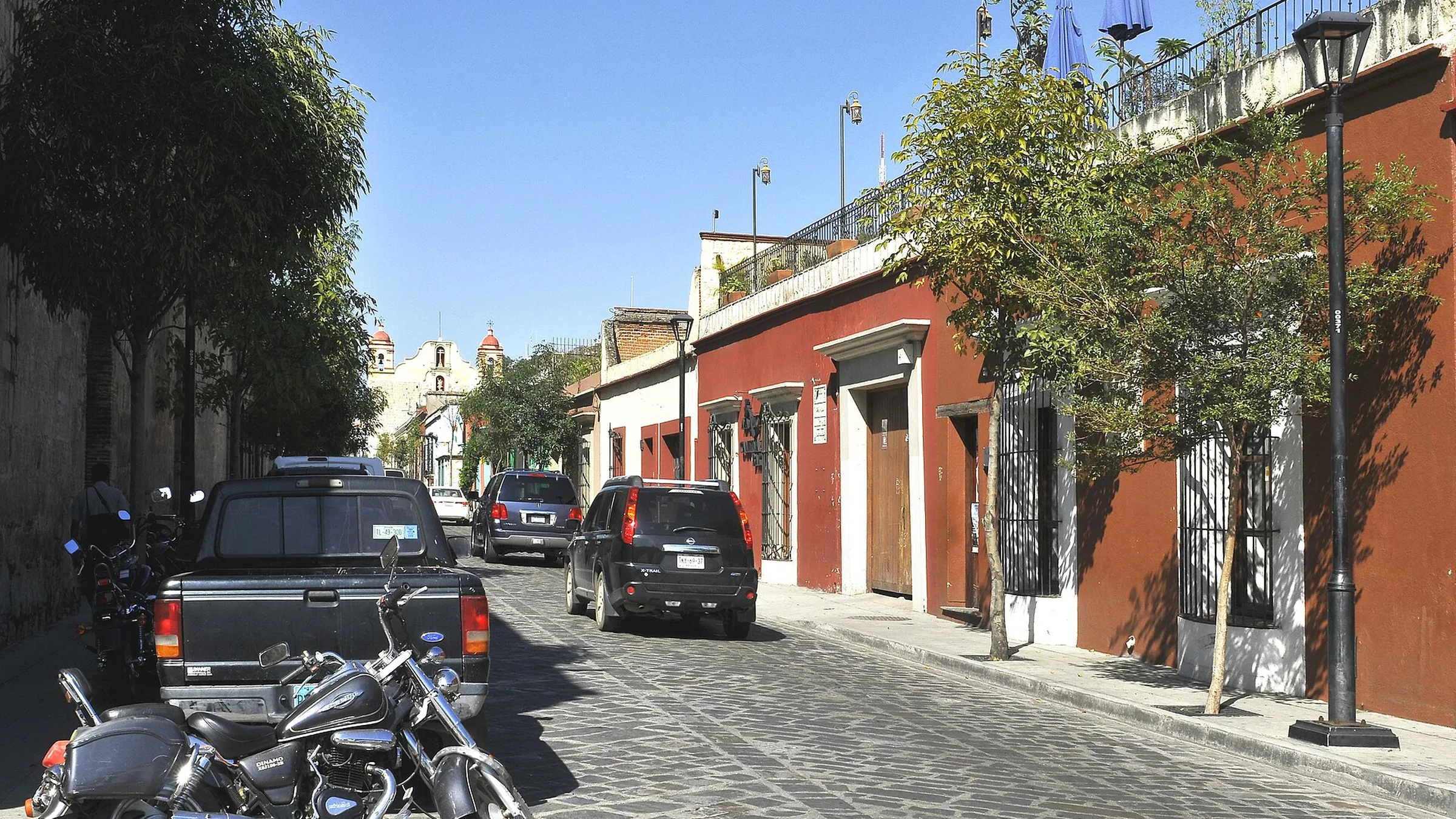 Calle empedrada del centro de Oaxaca de Juárez, con fachadas coloniales rojas y ocres, coches aparcados y una motocicleta en primer plano.