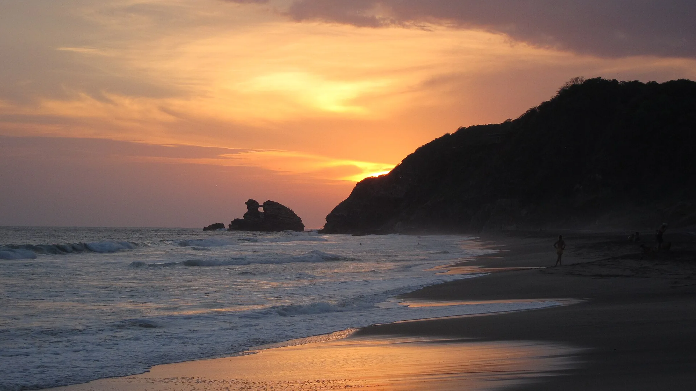 Playa Mermejita al atardecer, junto a Mazunte: el sol bajando detrás del cerro, las olas entrando en una arena larga y un islote rocoso recortado sobre el agua.
