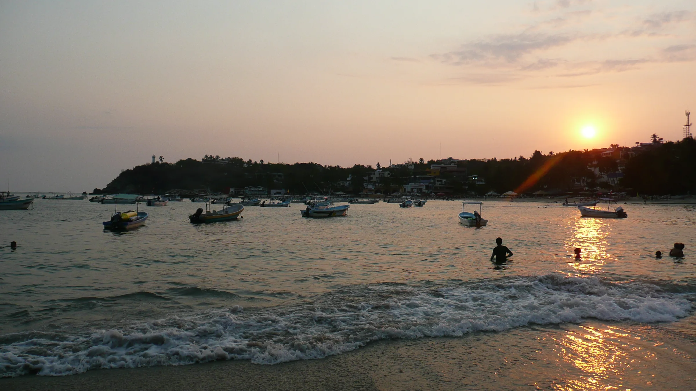 Bahía Principal de Puerto Escondido al atardecer: lanchas de pesca fondeadas en el agua, el pueblo y el faro sobre el promontorio y el sol bajando al fondo.