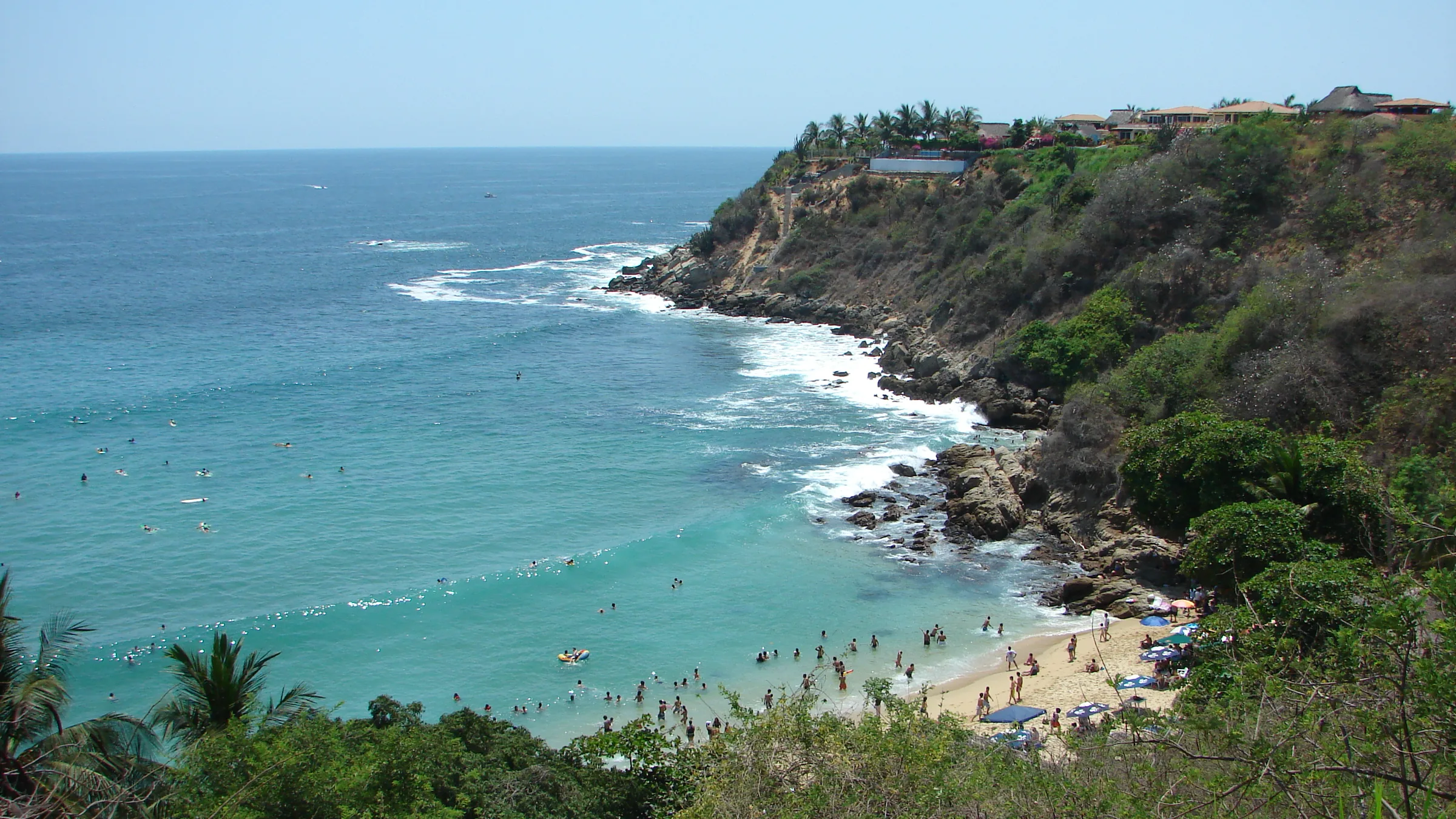 La cala de Carrizalillo en Puerto Escondido vista desde lo alto: agua turquesa con gente bañándose, una franja de arena al fondo y el acantilado verde con casas de tejado de palma en el borde.