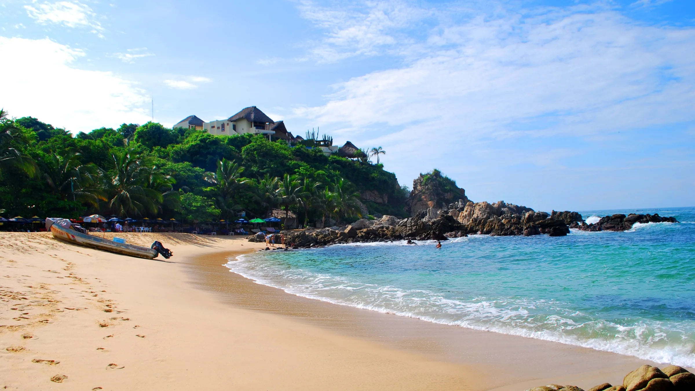 Playa Manzanillo en Puerto Escondido: una lancha de pesca varada en la arena, palmeras y palapas en la ladera y las rocas cerrando la cala sobre agua turquesa.
