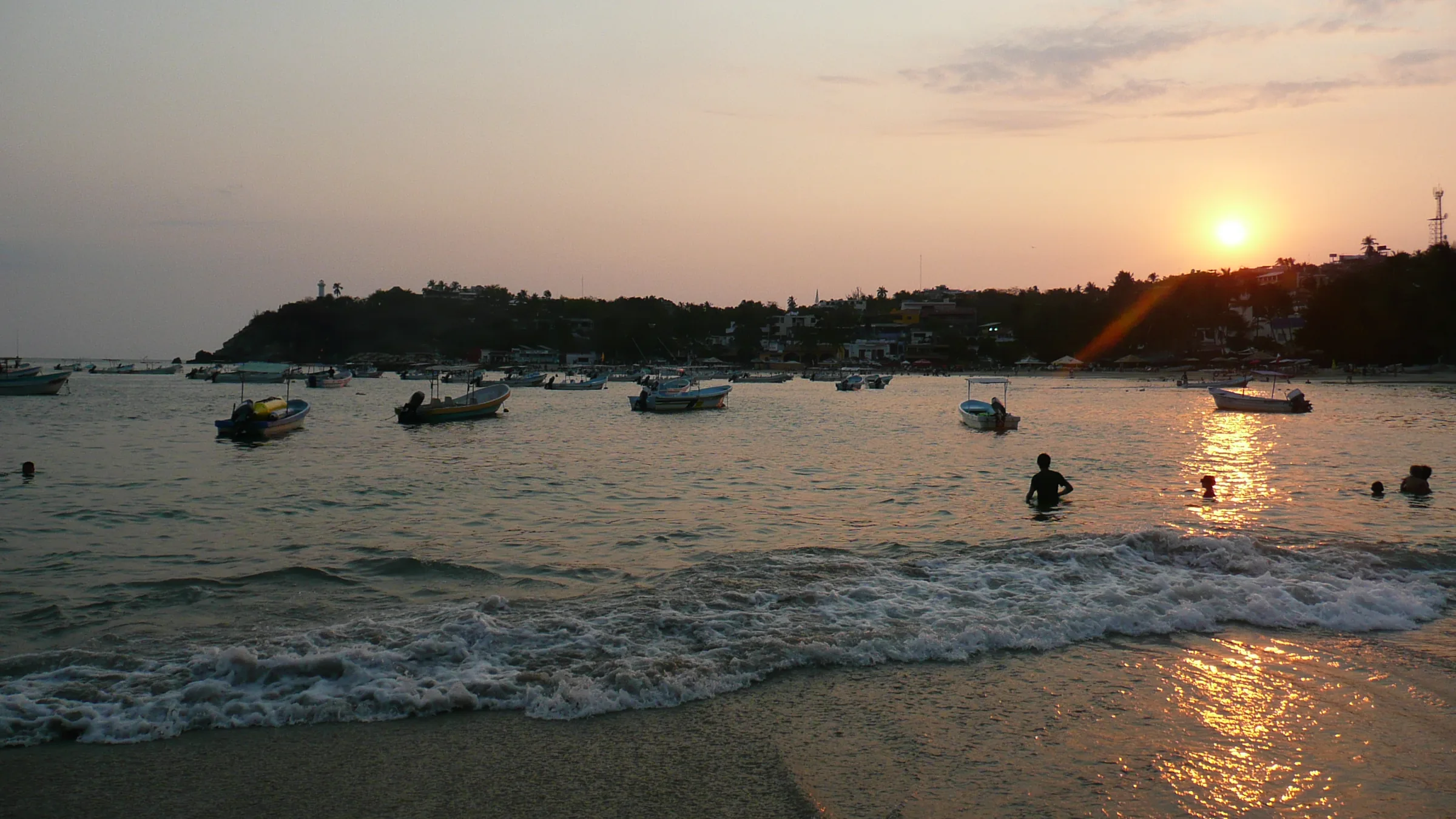 La Playa Principal de Puerto Escondido al atardecer: decenas de lanchas de pesca fondeadas en la bahía, bañistas en el agua a contraluz, el sol bajando sobre el cerro y el pueblo con su faro recortado en la punta.