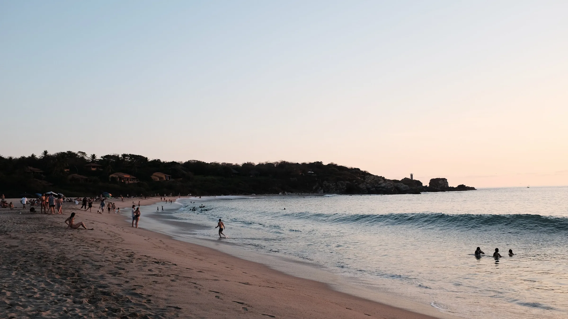 Punta Zicatela al atardecer: una playa larga de arena con el oleaje entrando en diagonal, un promontorio boscoso al fondo y el cielo rosado sobre el mar.