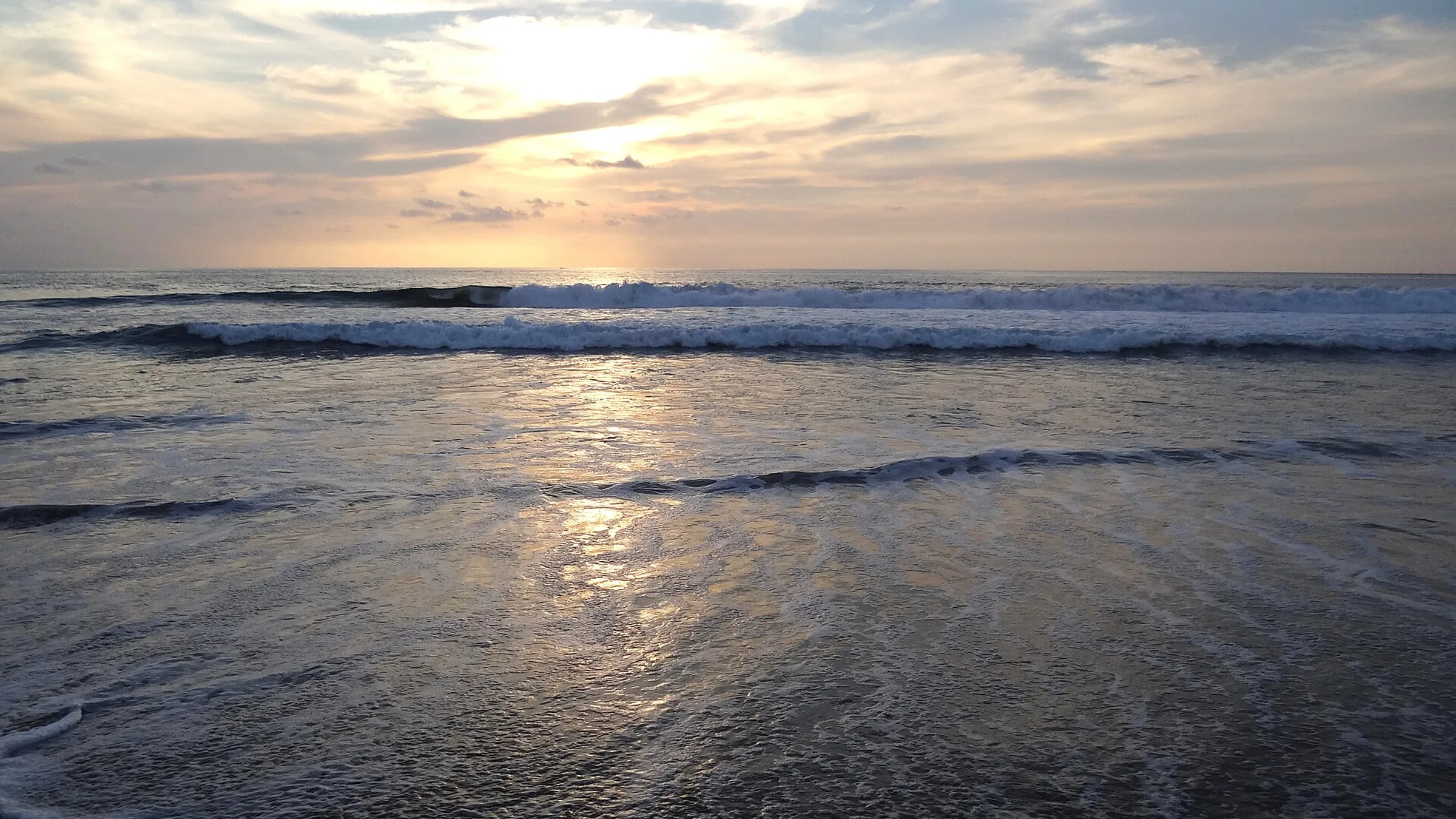 Atardecer en la playa de Zicatela, en Puerto Escondido: el sol bajo entre nubes sobre el mar, una serie de olas rompiendo en paralelo y el reflejo dorado sobre la arena mojada.