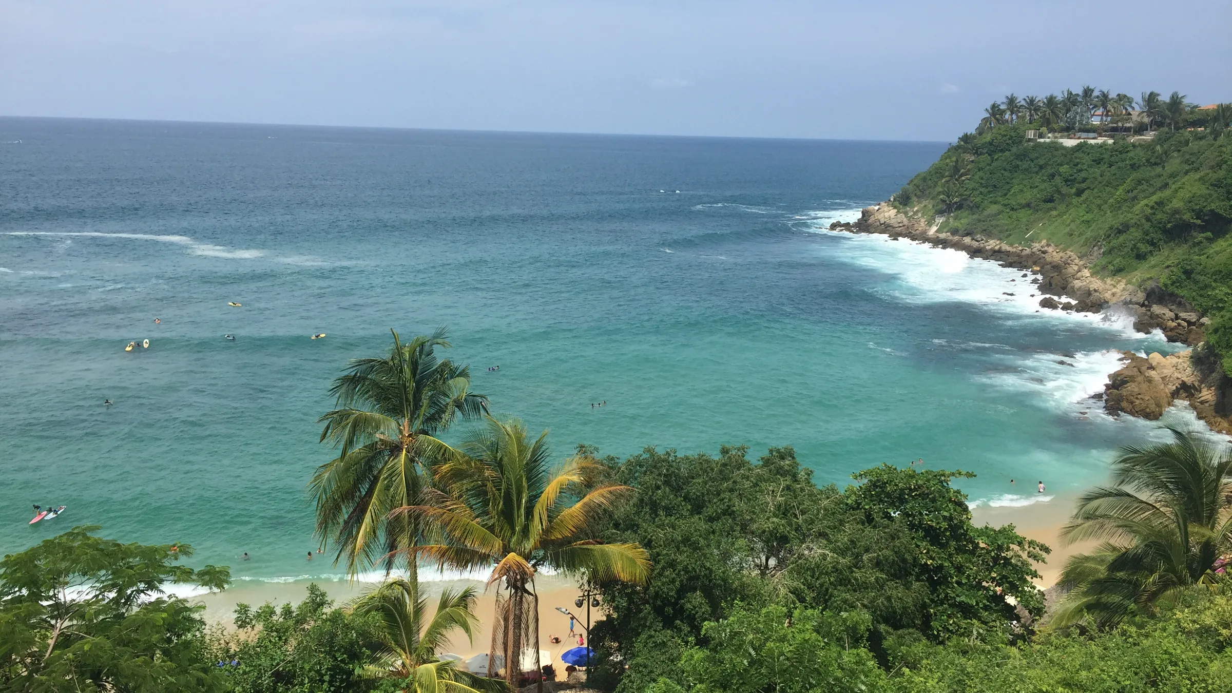 Carrizalillo visto desde arriba, desde el mirador del bulevar de Rinconada: las palapas y las palmeras del mirador en primer plano y, abajo, la cala con el agua turquesa, surfistas en el agua y la punta rocosa cerrando la bahía.