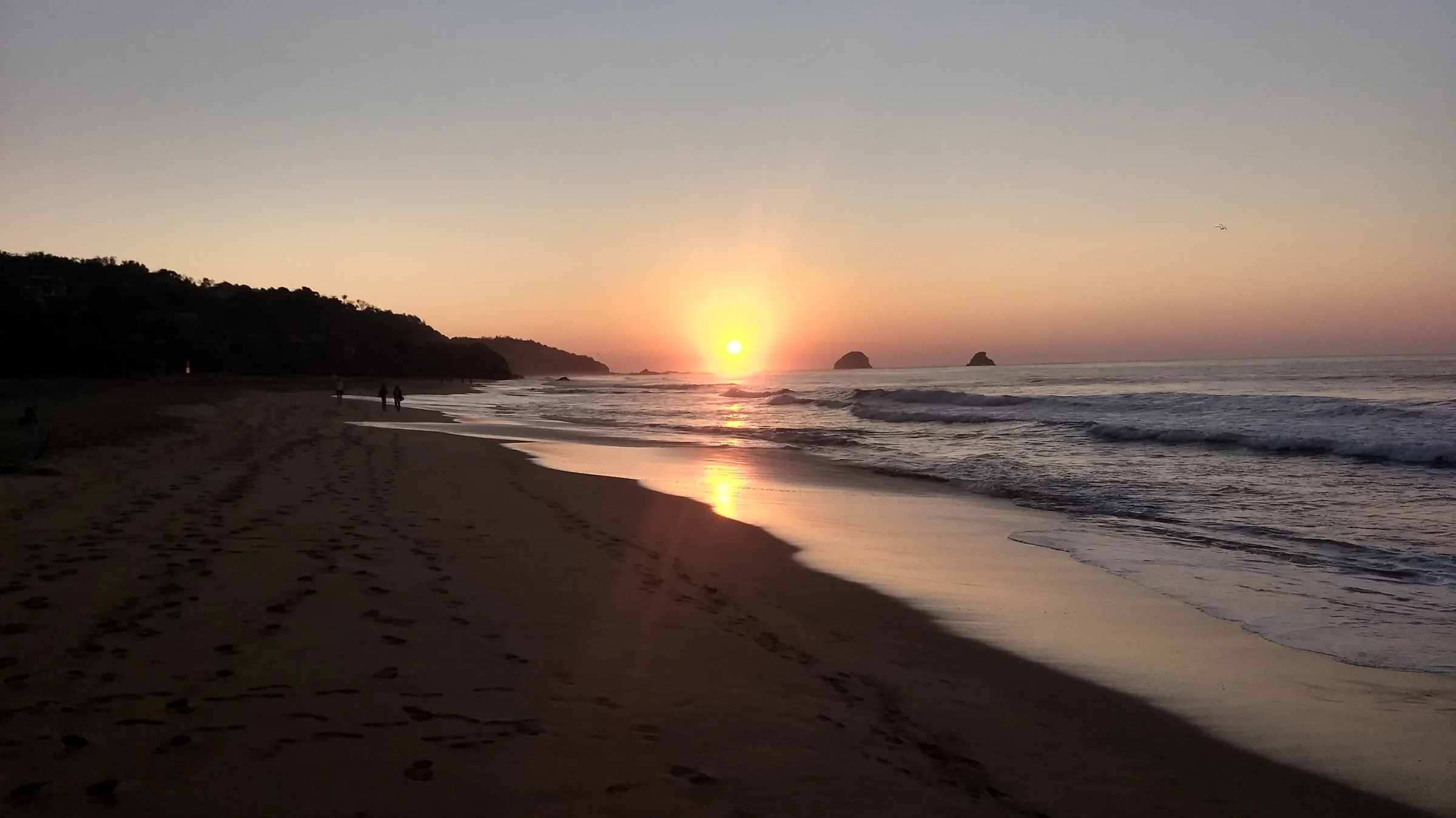 Atardecer en la playa de San Agustinillo: el sol bajando sobre el Pacífico, la arena húmeda reflejando la luz naranja, dos peñascos recortados en el horizonte y unas pocas personas caminando por la orilla.