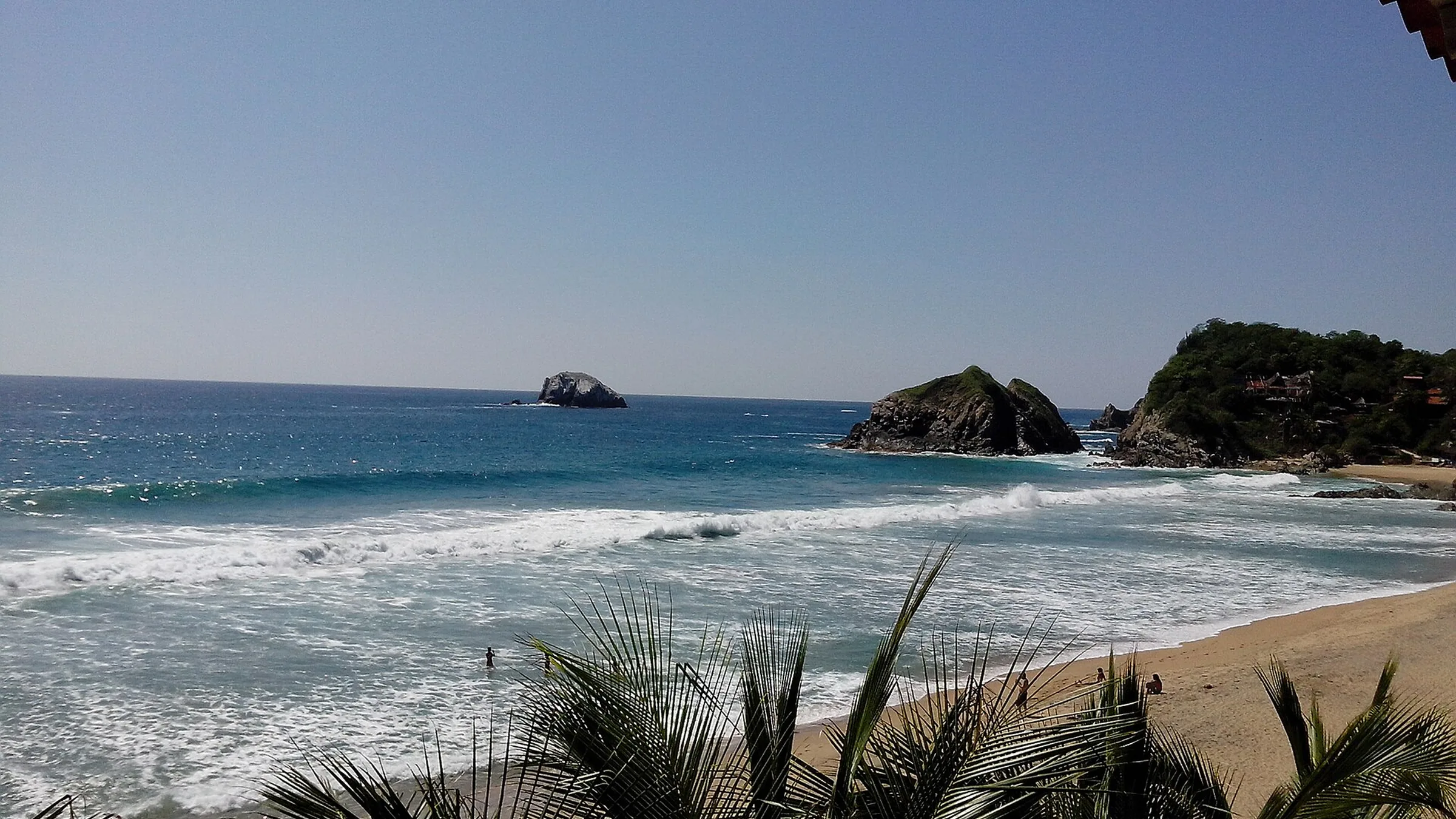 Playa de Zipolite vista entre palmeras: olas rompiendo en una arena larga y dos islotes rocosos frente a la costa.